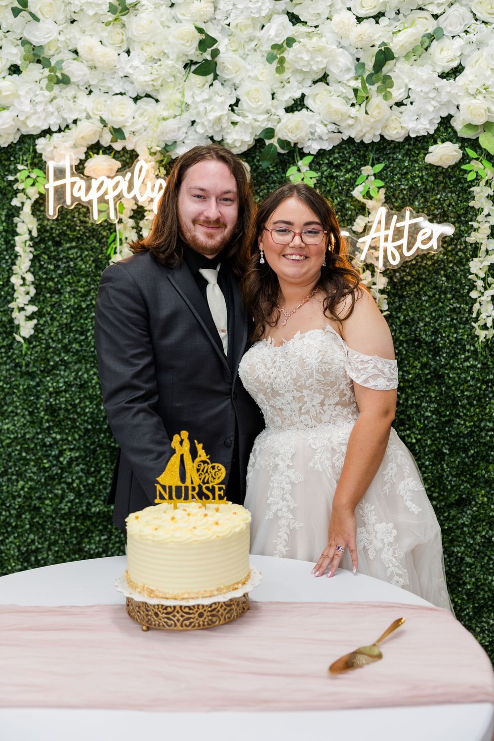 Newlywed couple posing with wedding cake in front of floral wall with Happily Ever After sign.
