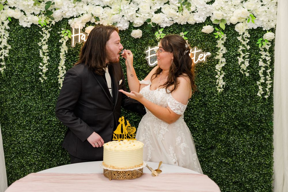 Bride and groom sharing cake at wedding reception in front of floral green wall backdrop.
