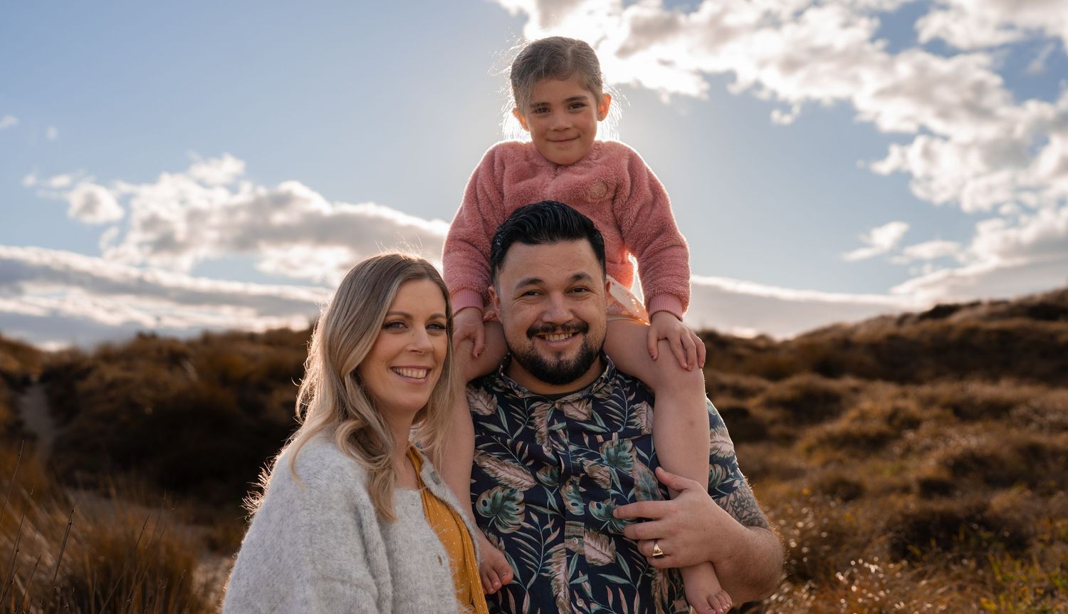 Happy family of three posing outdoors, parents smiling with toddler on dad's shoulders against a sunny sky.