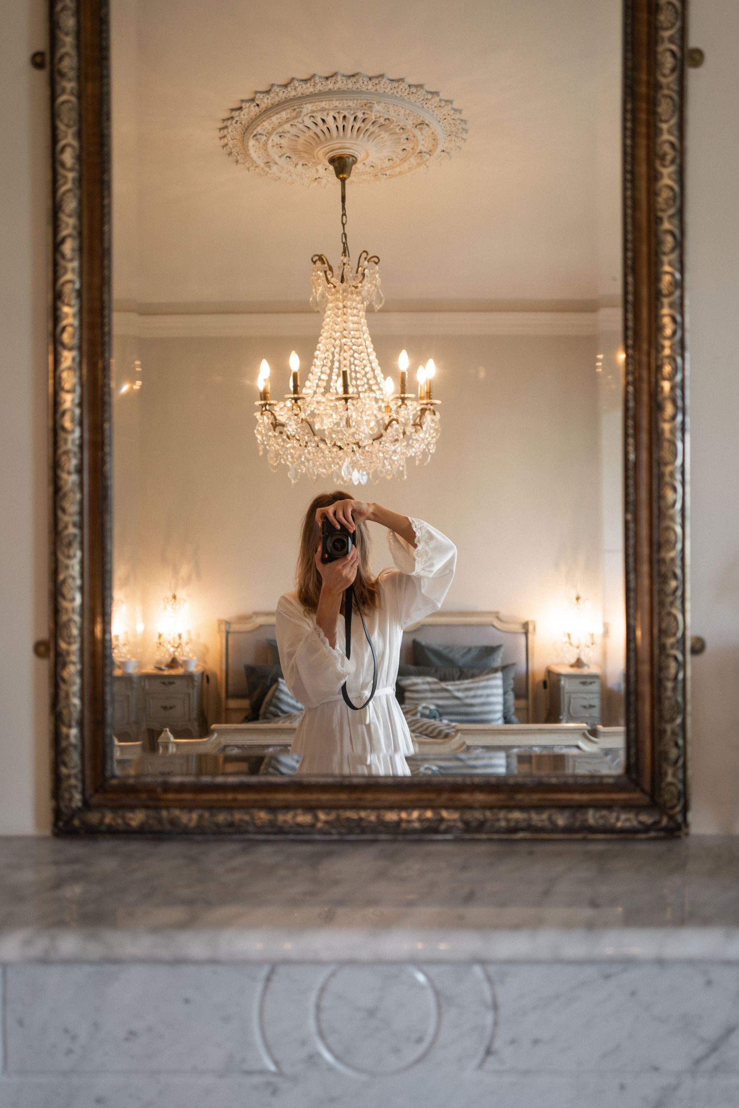 Woman in white robe taking mirror selfie in elegant bedroom with crystal chandelier and ornate gold frame.