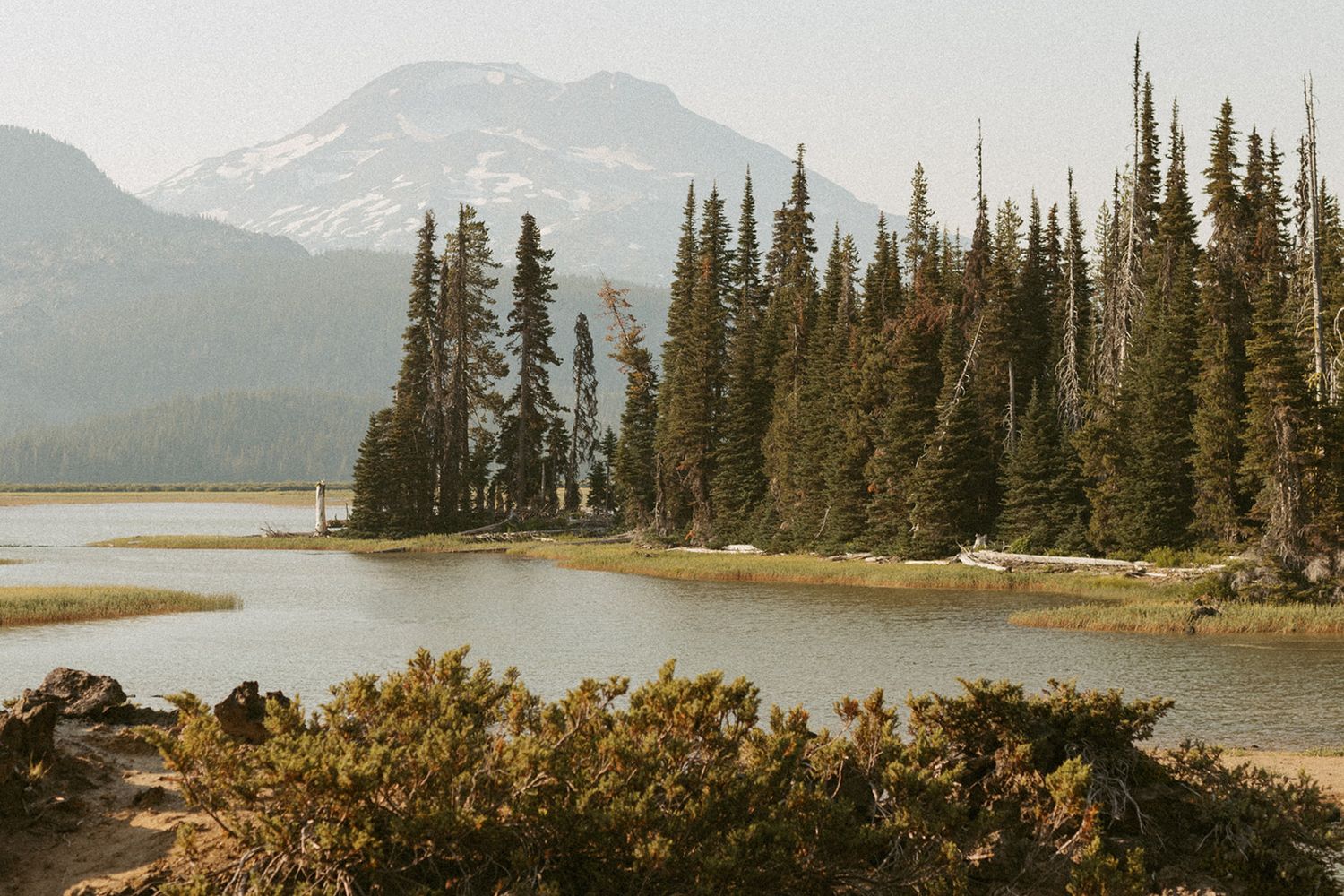 Serene mountain lake surrounded by tall evergreen trees with a snow-capped peak in the background.