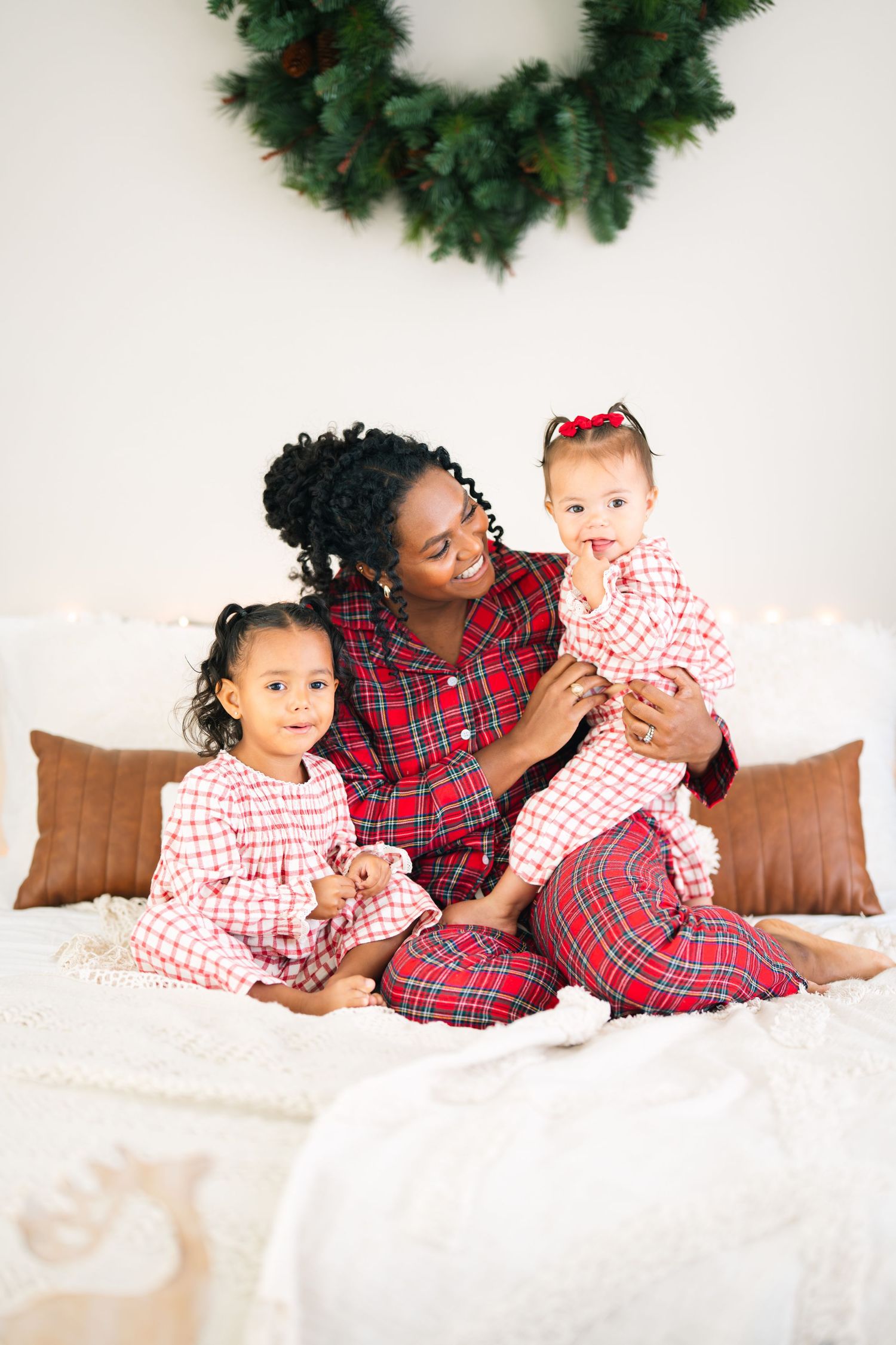 A mother and two young daughters in matching red plaid Christmas pajamas sit on a bed with a festive wreath backdrop.