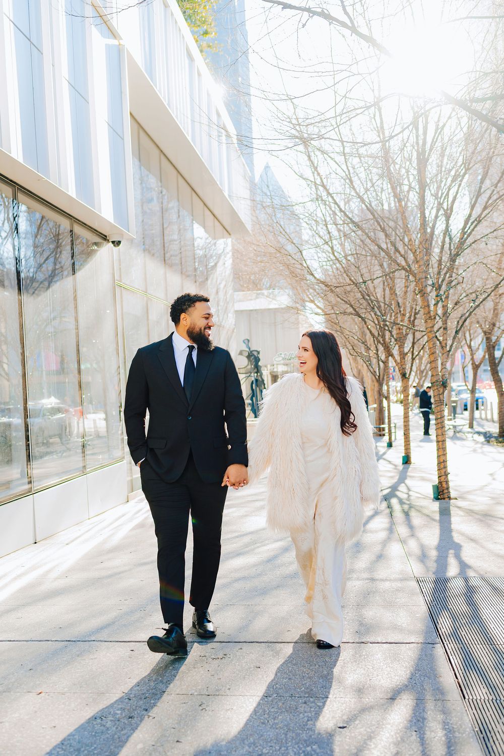 A couple walks hand-in-hand on a sunny urban sidewalk, man in black suit, woman in white outfit, smiling at each other.