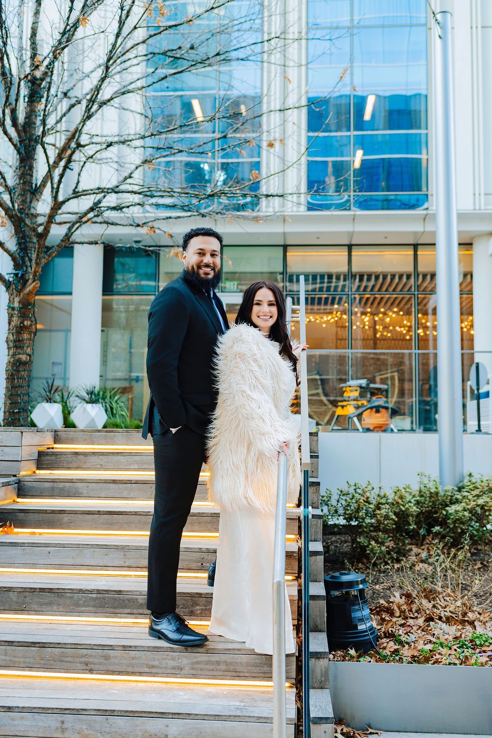 Elegant couple posing on outdoor steps, woman in white dress and fur coat, man in black suit, modern building backdrop.
