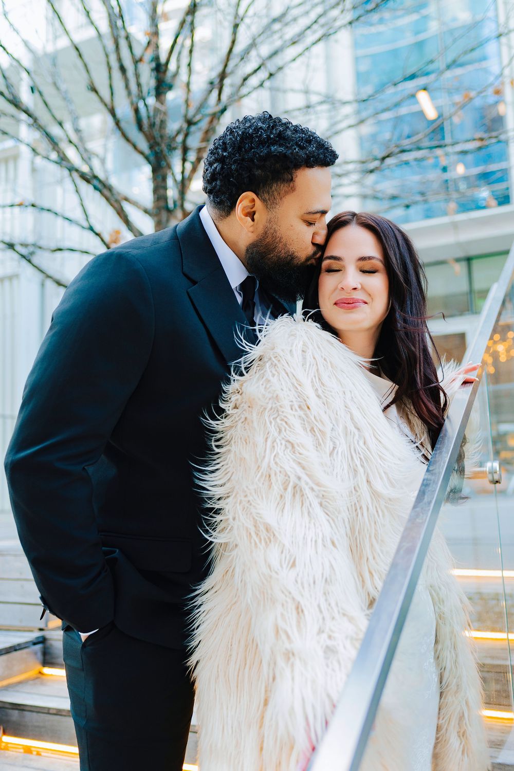 A man in a black suit kisses a woman in a fluffy white coat on the cheek outdoors in a city setting.