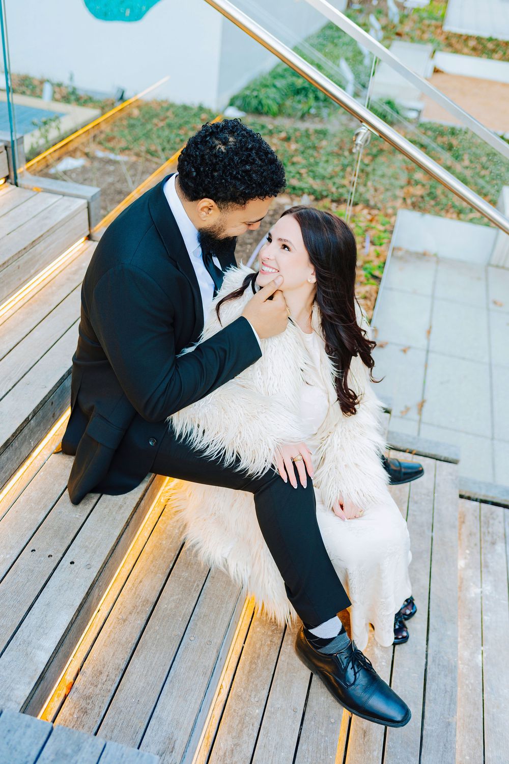 Romantic couple sitting on wooden steps, woman in white fur coat smiling at man in black suit.