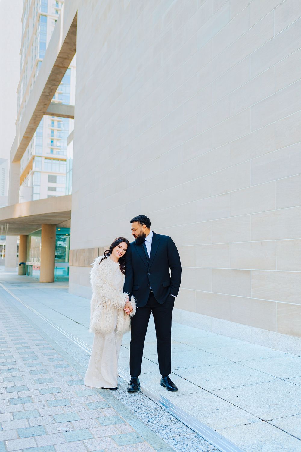 Couple in formal attire poses outside modern building, woman in white fur coat, man in black suit holding hands.