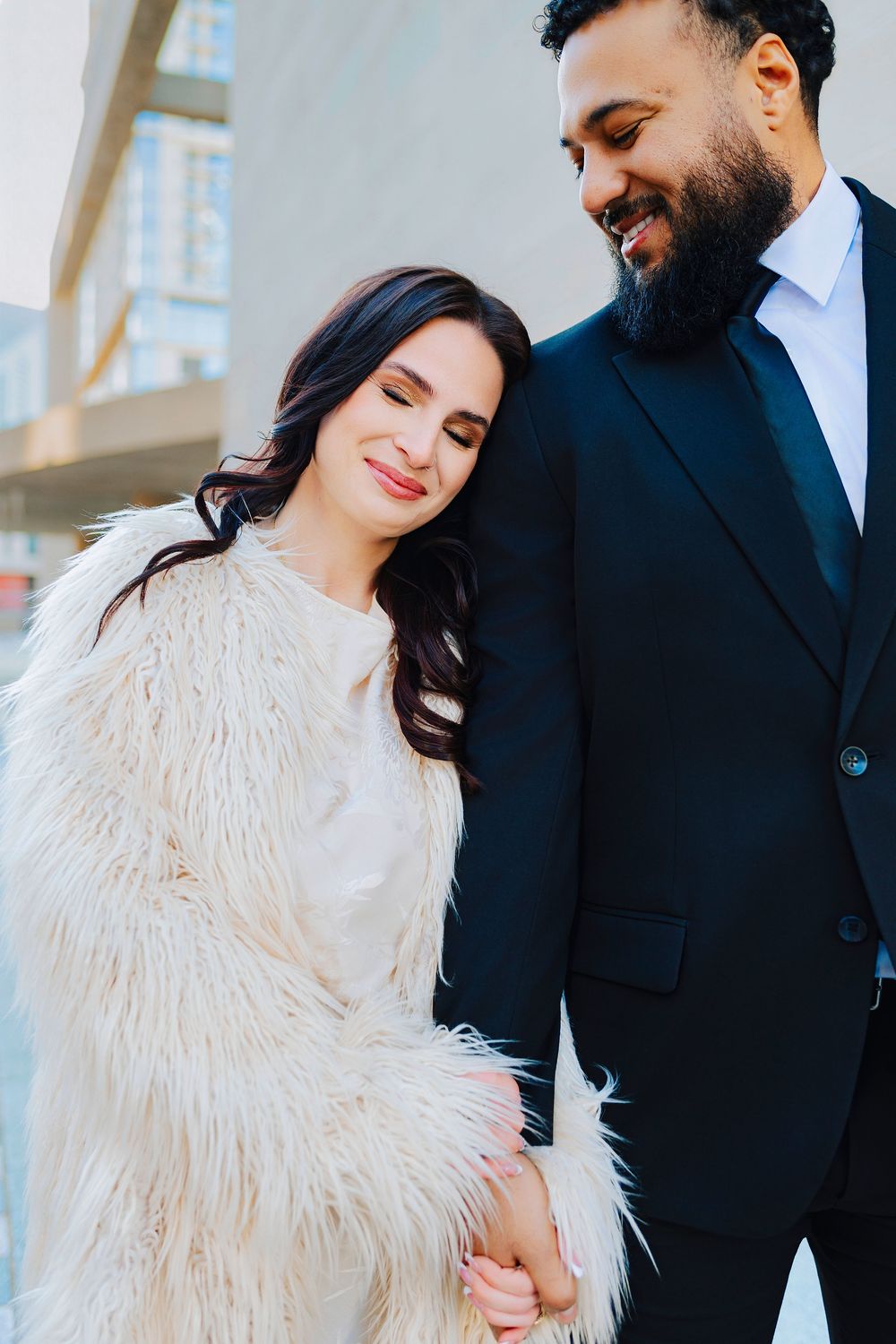 A smiling couple in elegant attire, woman in white fur coat leans on bearded man in black suit outdoors.