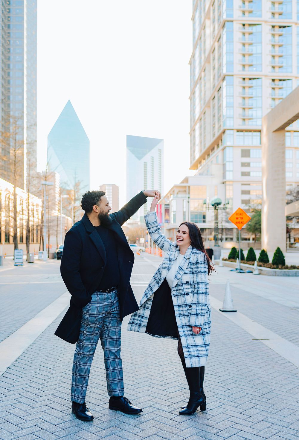 Couple dancing outdoors in Dallas cityscape, wearing stylish plaid coats, holding hands raised in urban setting.