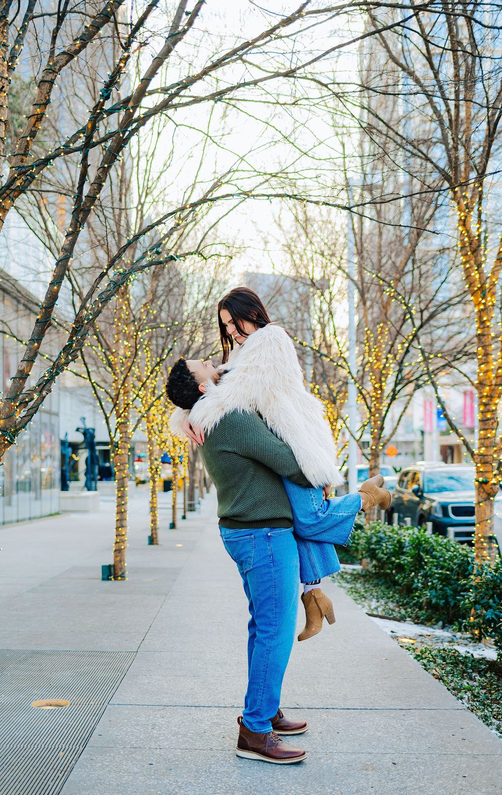 Man lifting woman among trees with twinkling lights on a city sidewalk during winter engagement photo session.