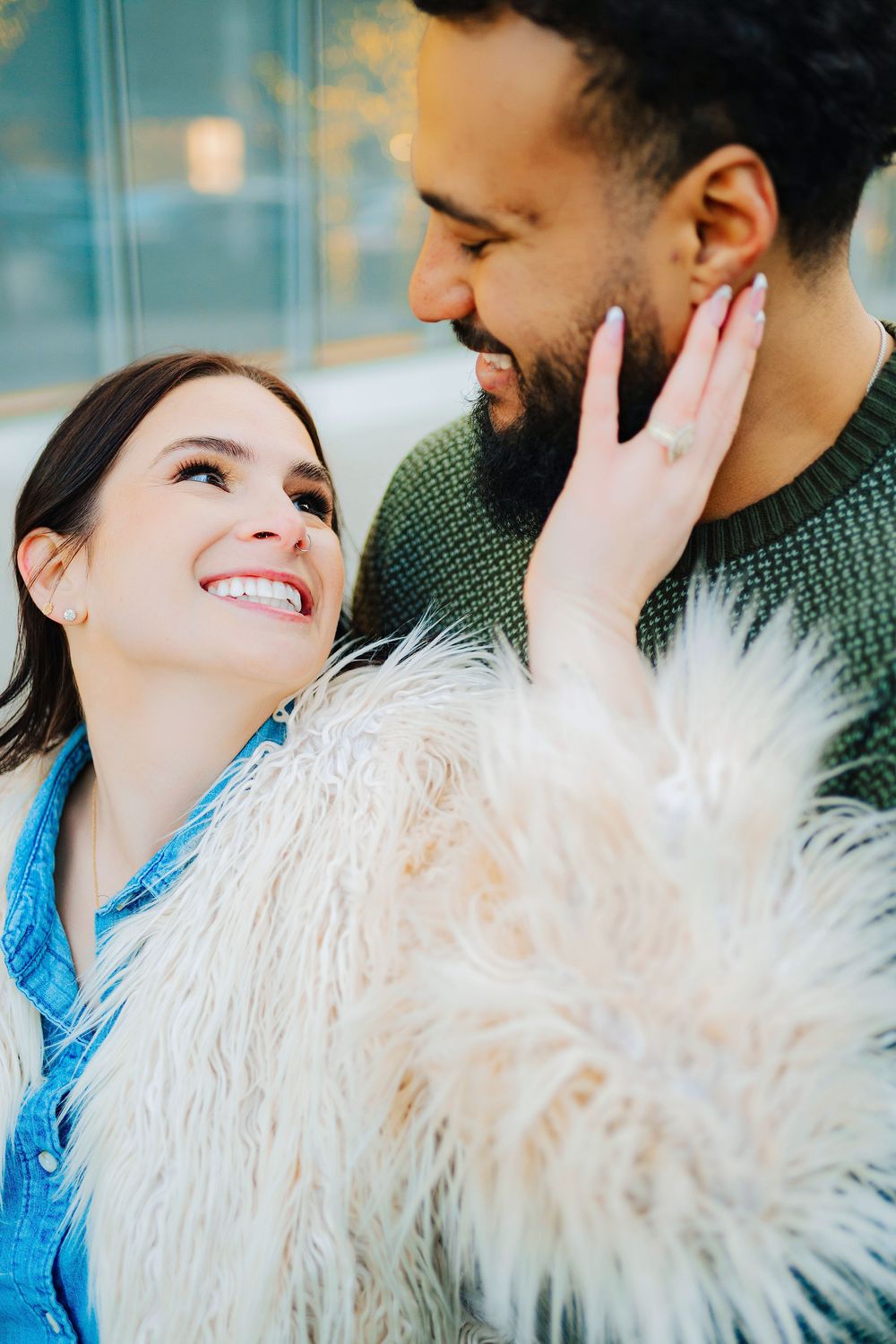 A smiling woman in a faux fur coat tenderly touches a bearded man's face as they share a romantic moment.