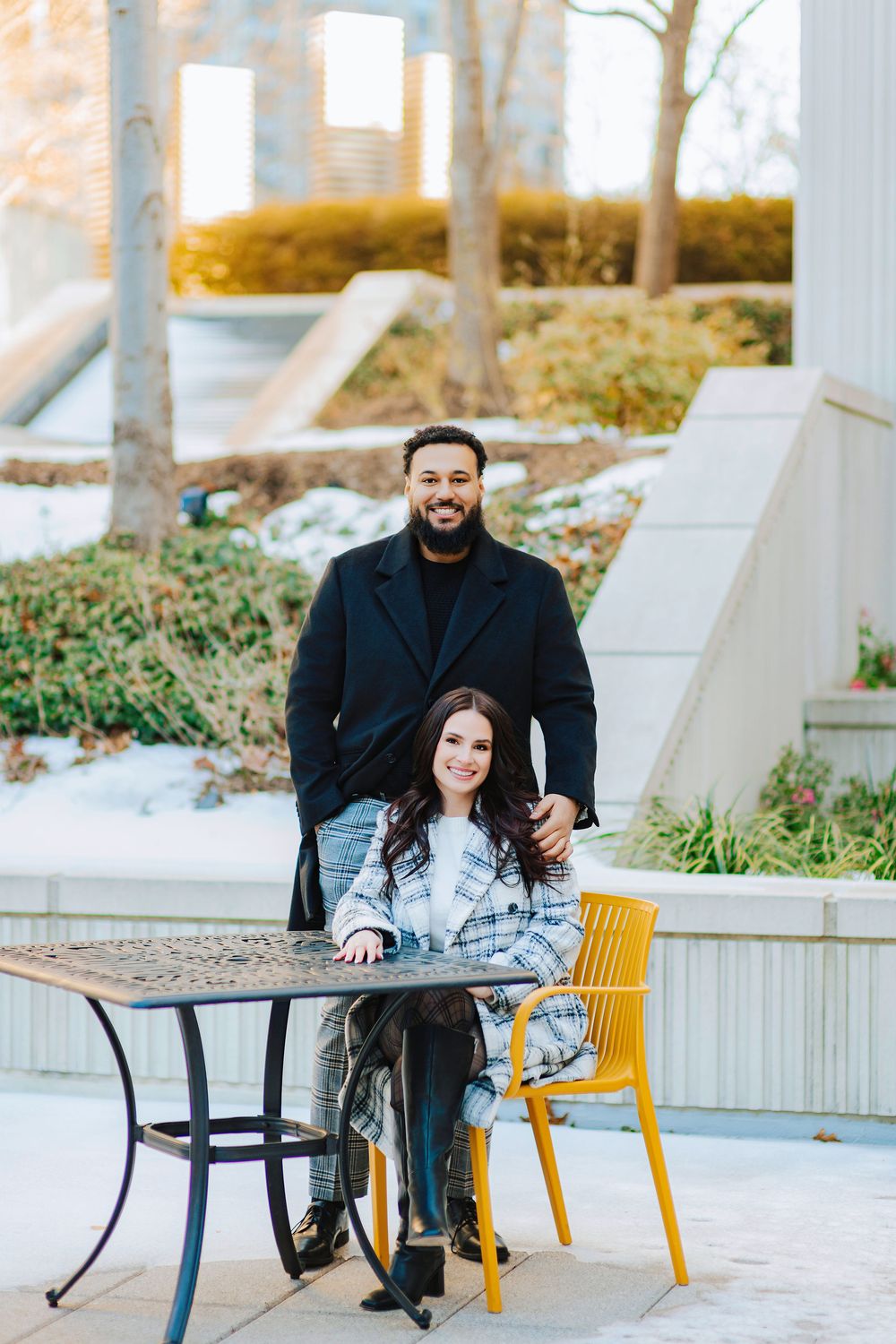 A smiling couple poses outdoors at a metal table, man in black coat standing behind seated woman in plaid jacket.