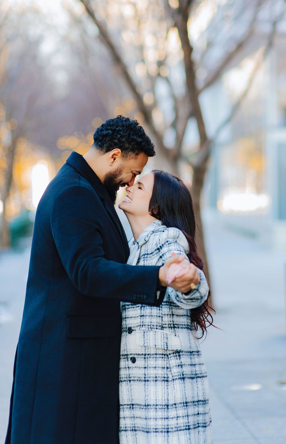 A couple in winter coats shares a romantic embrace on a tree-lined street during golden hour.