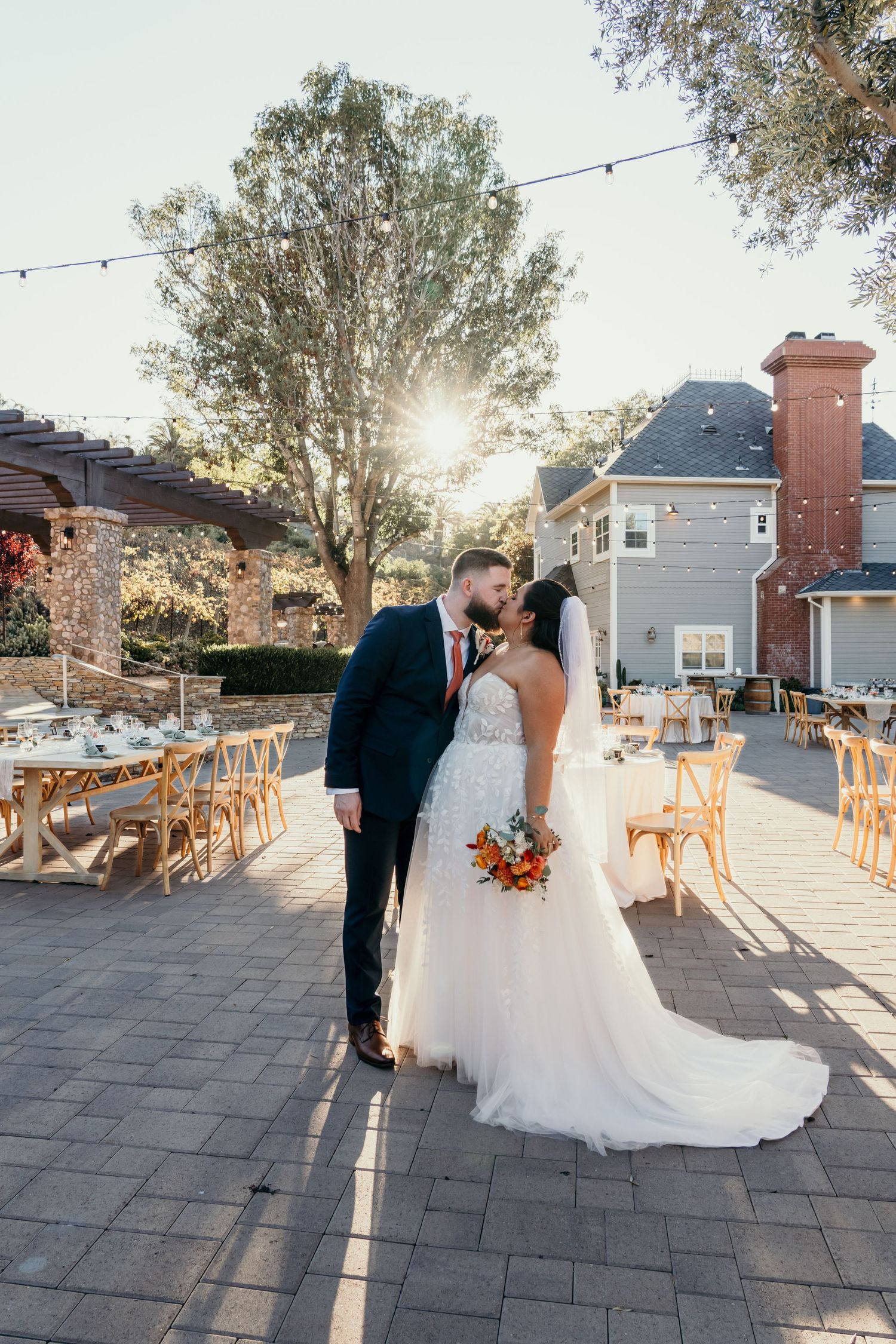 Bride and groom share a kiss at golden hour on a patio wedding venue with string lights and reception tables.