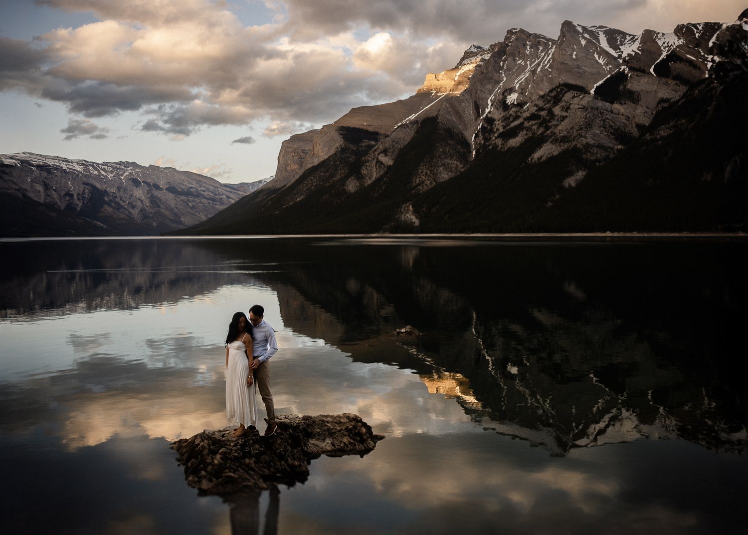 Couple embracing on a rock in a mirror-calm mountain lake at sunset, by a Banff wedding photographer
