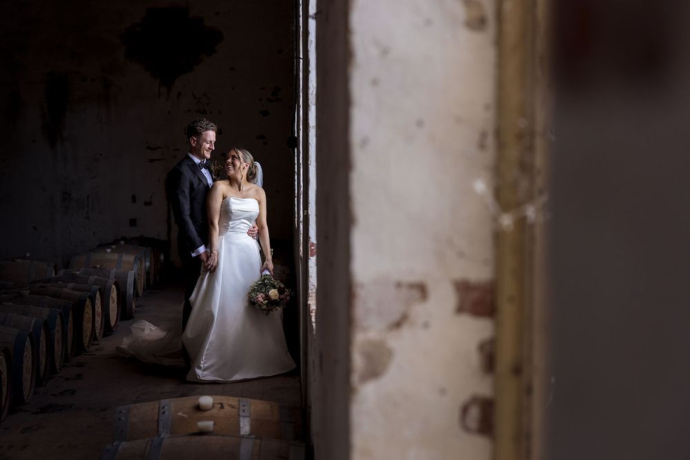 Bride in a white gown holding a bouquet embraces her groom in a dark suit inside a rustic cellar with aged walls and wooden barrels.