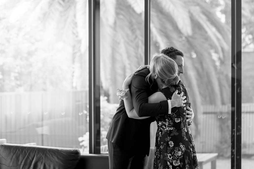 Mother in a floral dress pins a boutonniere on a groom in a black suit, then fixes his lapel flower in a bright room with large windows.