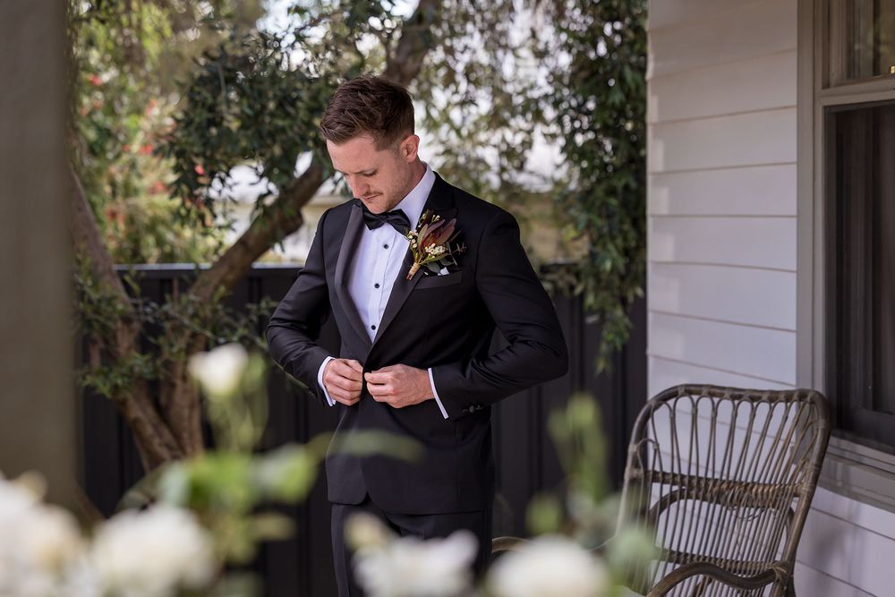 Groom in a black tuxedo with bow tie and floral boutonniere adjusts his jacket on a garden porch beside a wicker chair.