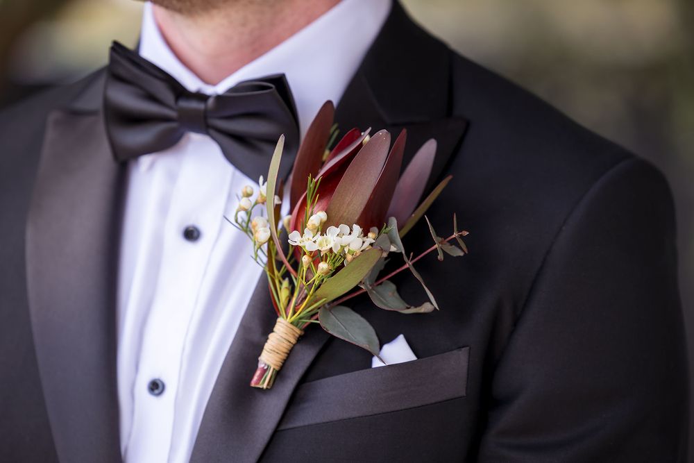 Groom in a black tuxedo with a bow tie wearing a rustic boutonniere of burgundy leaves, white flowers and greenery wrapped with twine.