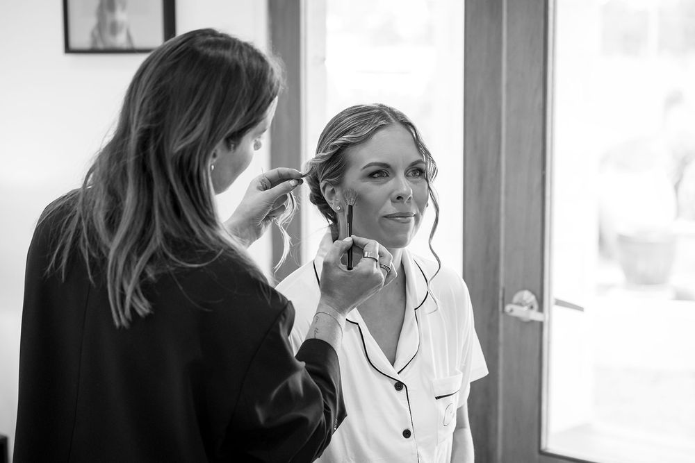 A makeup artist pins and styles a smiling woman's updo near a bright window in this black and white getting-ready photo.