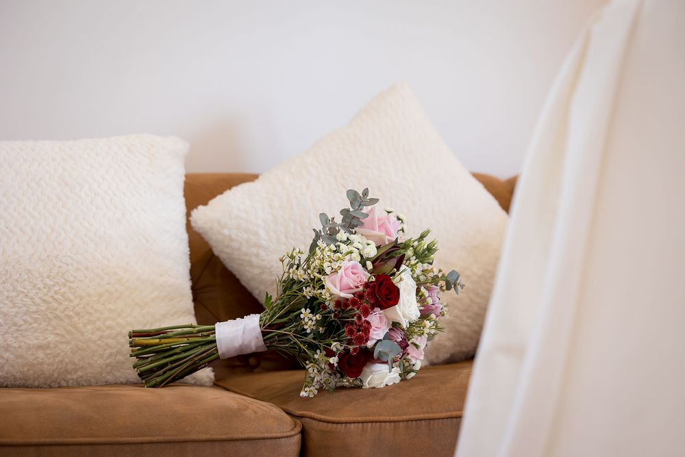 Pink, burgundy and white wedding bouquet wrapped in a white ribbon resting on a tan leather sofa beside cream textured throw pillows.