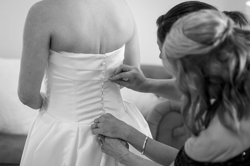 Bridesmaid with her hair pulled back carefully buttons the back of a strapless white wedding gown in a black and white photo.