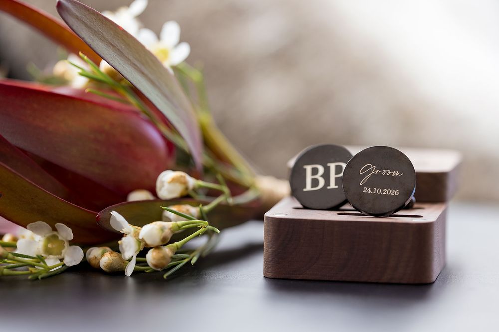 Personalized round engraved cufflinks with initials and a wedding date resting on a walnut wooden box beside delicate white flowers and burg