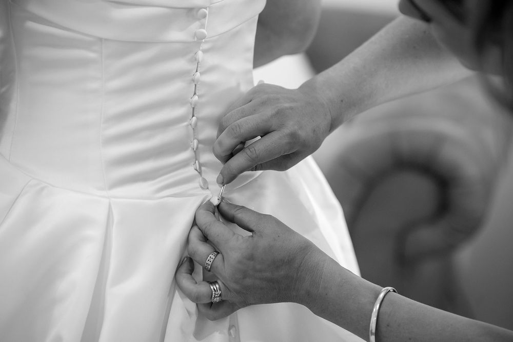 Black and white photo of two hands fastening the buttons on the back of a satin wedding dress before the ceremony.