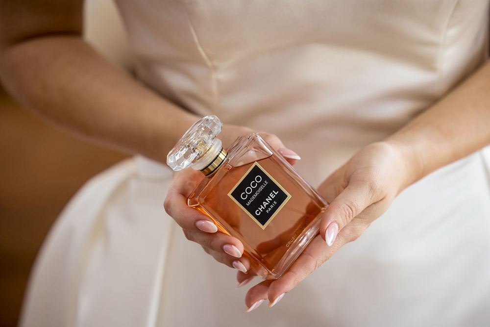 Bride in her wedding dress holding a bottle of Coco Chanel perfume with amber liquid and a clear faceted cap in her manicured hands.