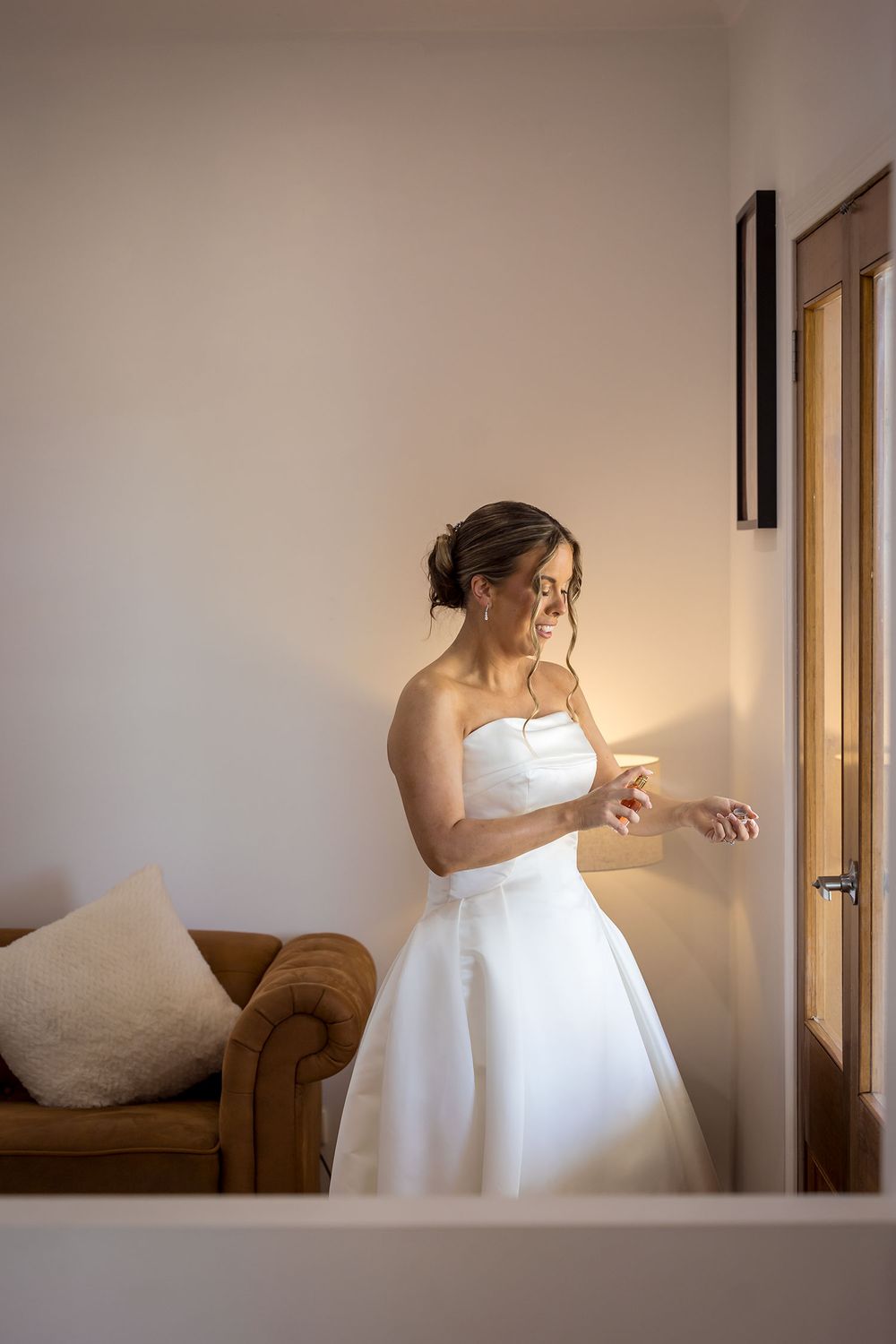 Bride in a strapless white satin wedding gown spays perfume while getting ready in a softly lit room beside a tan leather armchair.