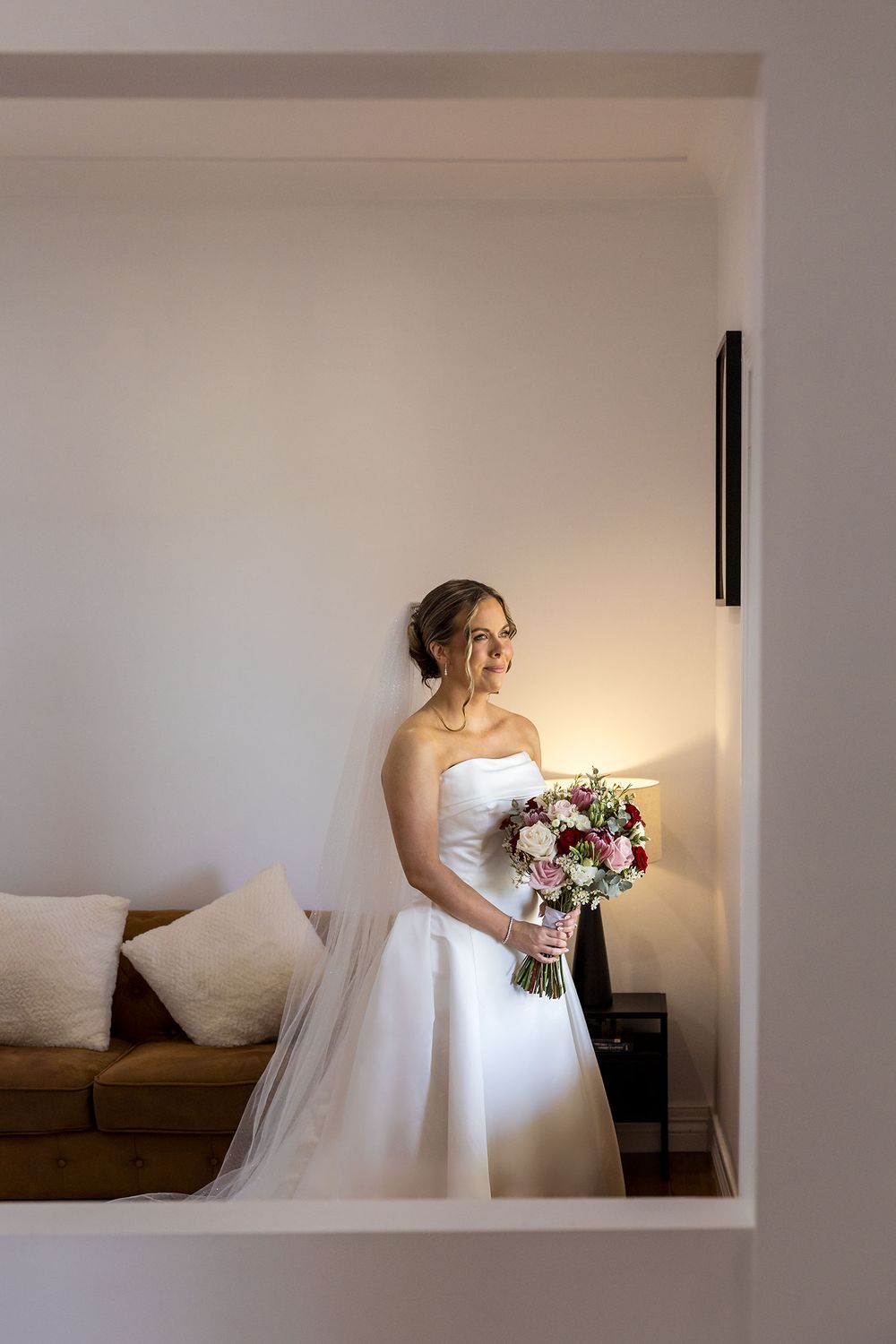 Bride in a strapless white gown and long veil holds a pink and white bouquet while gazing out a window beside a cozy sofa.
