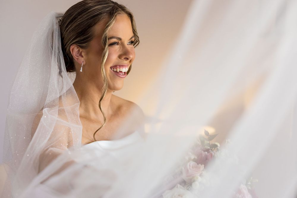 Smiling bride in a white wedding dress and sheer veil with pearl earrings, holding a pink floral bouquet in soft natural window light.