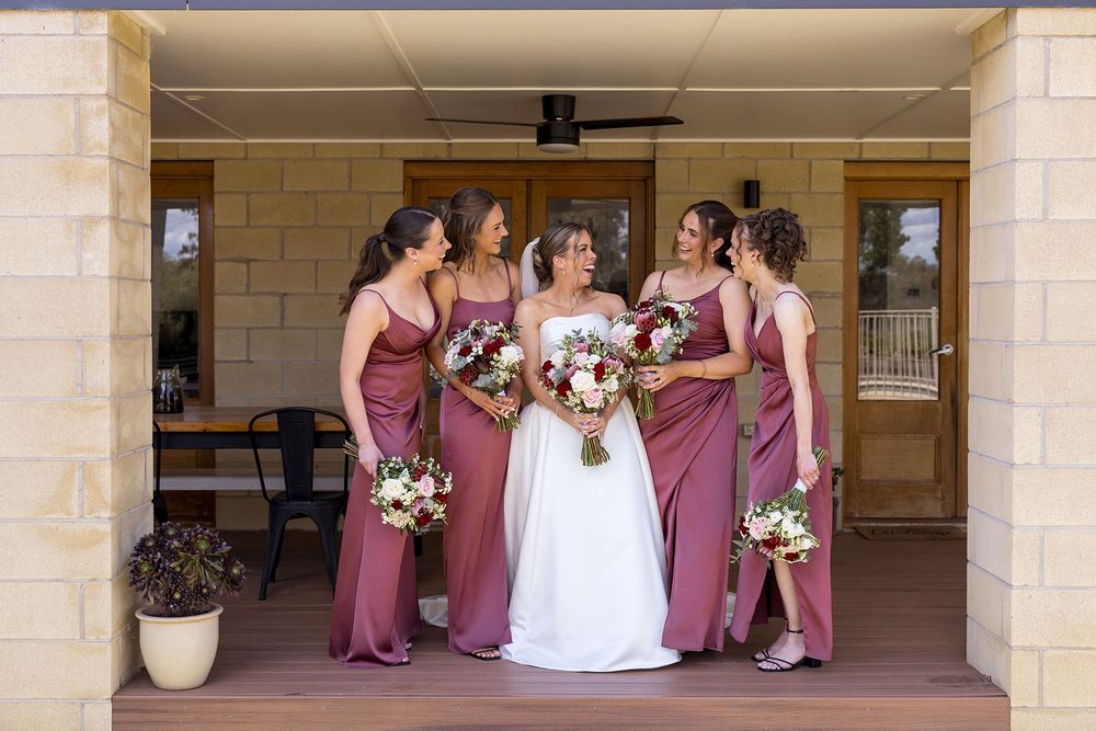 Bride in a white wedding gown holding a bouquet, standing with five bridesmaids in dusty rose dresses on a rustic stone porch.