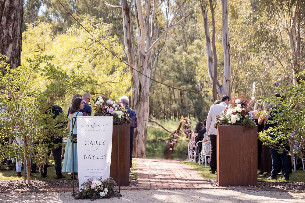 Wedding guests gather along a brick aisle lined with floral arrangements and a Carly and Bayley welcome sign in a sunlit eucalyptus forest.