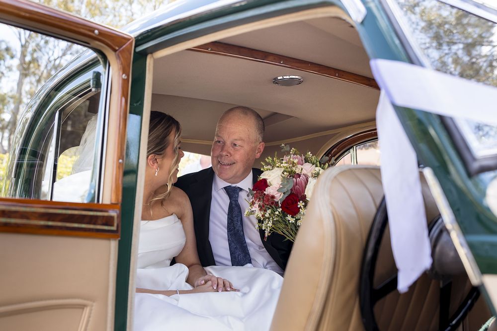 A bride in a white gown holding a bouquet sits beside her Dad in a suit in the back of a vintage green wedding car on wedding day.