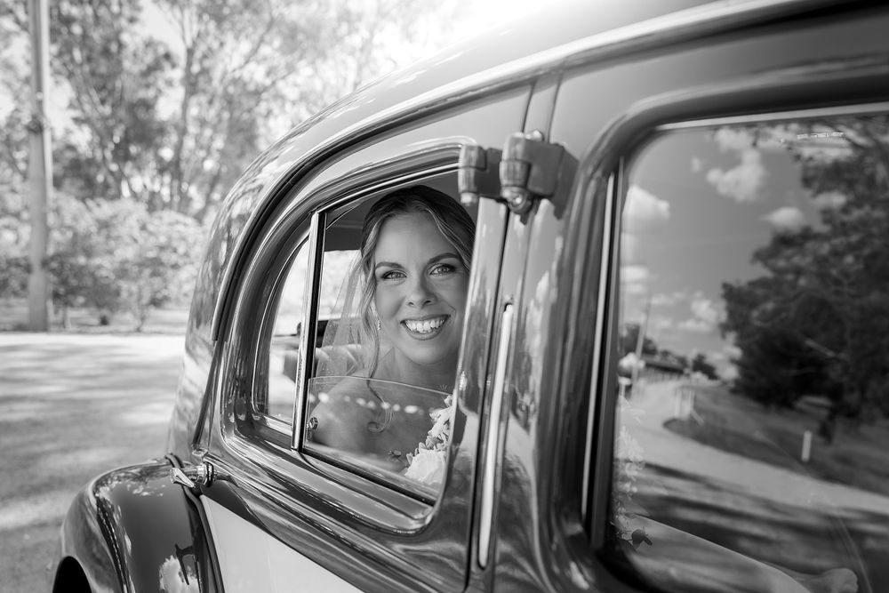 Black and white photo of a smiling Bride in a pearl necklace looking out the window of a vintage car, with trees reflected in the glass.