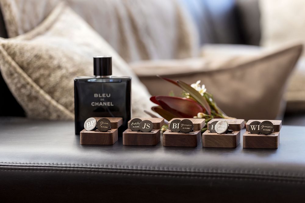 Bleu de Chanel cologne bottle beside a bouquet and four wooden monogrammed cufflink boxes on a leather bench with cushions.