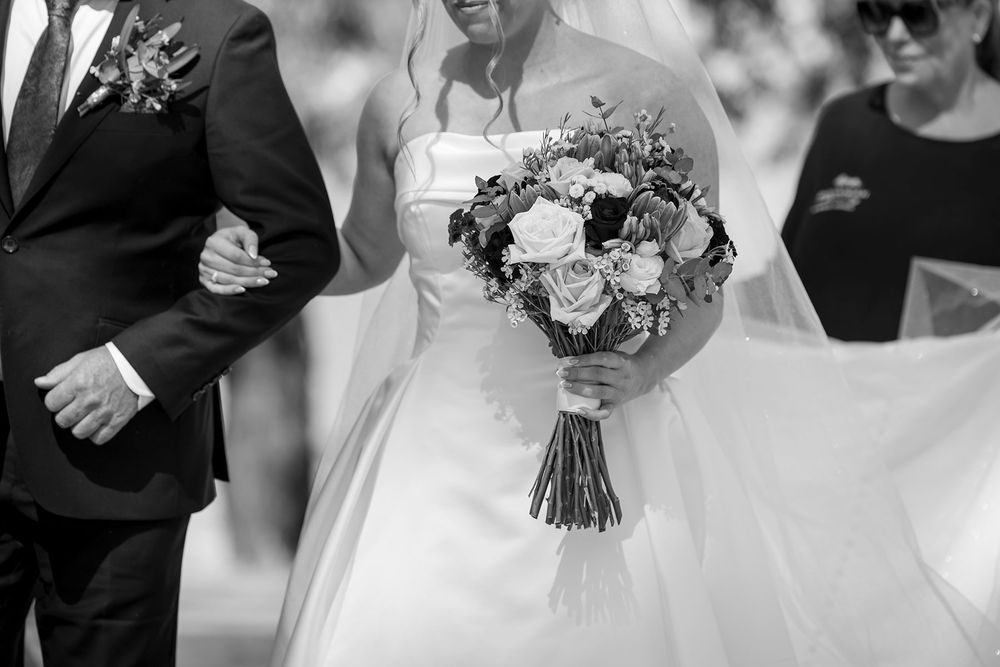 Black and white photo of a bride in a strapless gown holding a rose bouquet while walking arm in arm with her Dad in a suit.