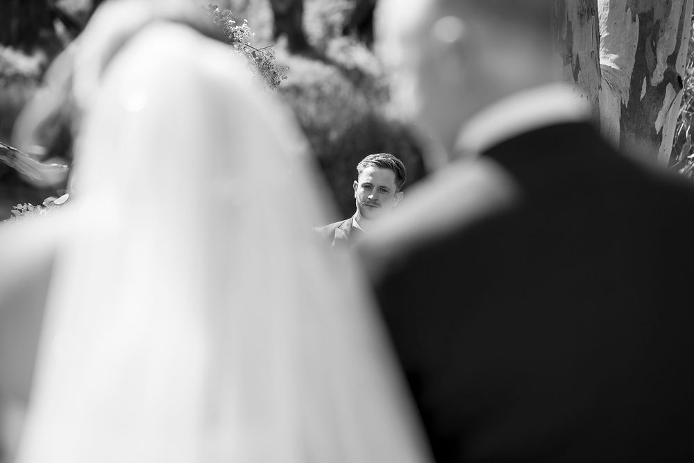 Black and white wedding photo of a groom smiling at his approaching bride, framed by the blurred veil and father of the bride.