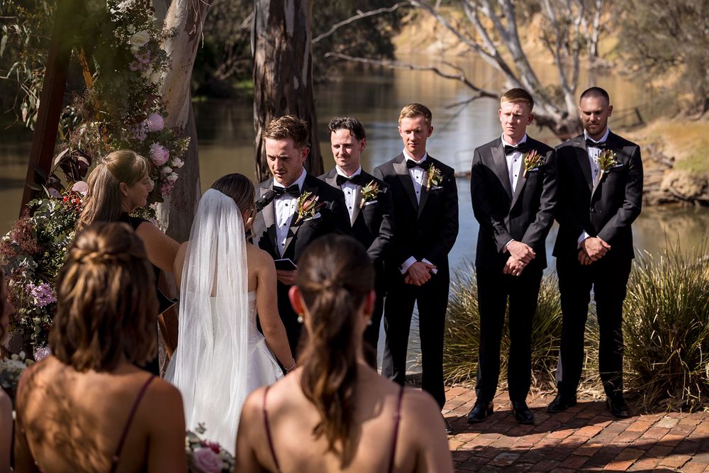 Groomsmen in dark suits with floral boutonnieres stand beside a bride in a white veil during an outdoor riverside wedding ceremony.