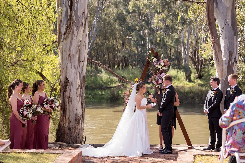 Bride in a white gown and groom exchange vows at an outdoor riverside wedding ceremony with bridesmaids in burgundy and groomsmen watching.