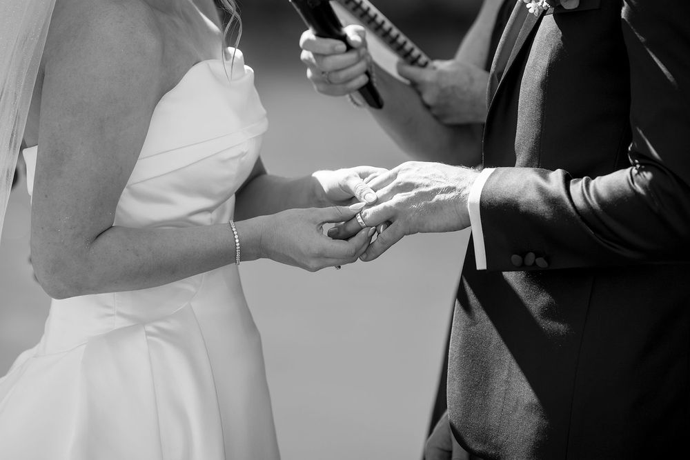 Black and white photo of a bride in a strapless gown and groom in a suit exchanging rings during a wedding ceremony.