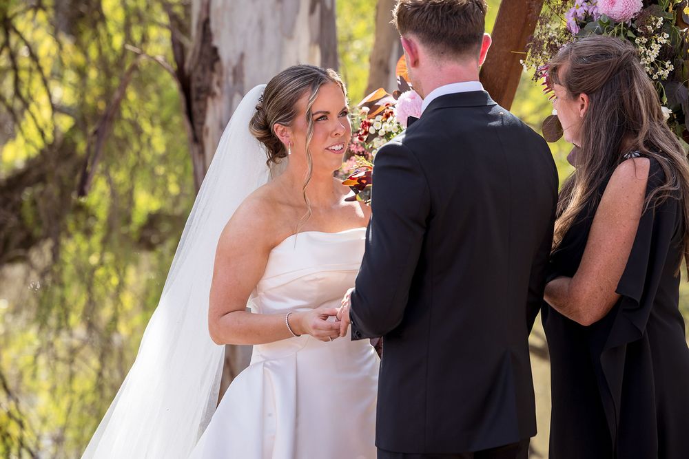 Smiling bride in a strapless white gown and long veil holds hands with the groom during an outdoor wedding ceremony beneath willow trees.