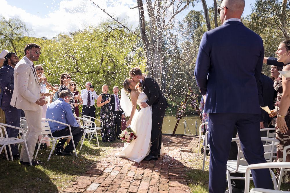 Bride and groom kiss at an outdoor garden wedding ceremony as guests seated in white chairs watch on a brick path.