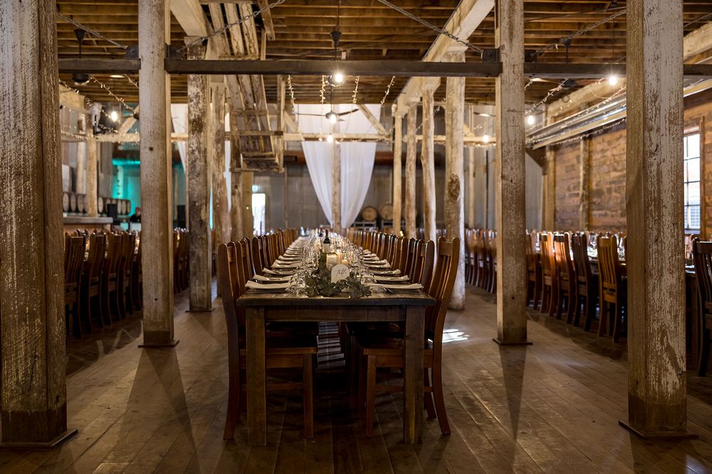 Long rustic wooden banquet table set for an event in a spacious converted warehouse venue with exposed beams, tall columns, and brick walls.