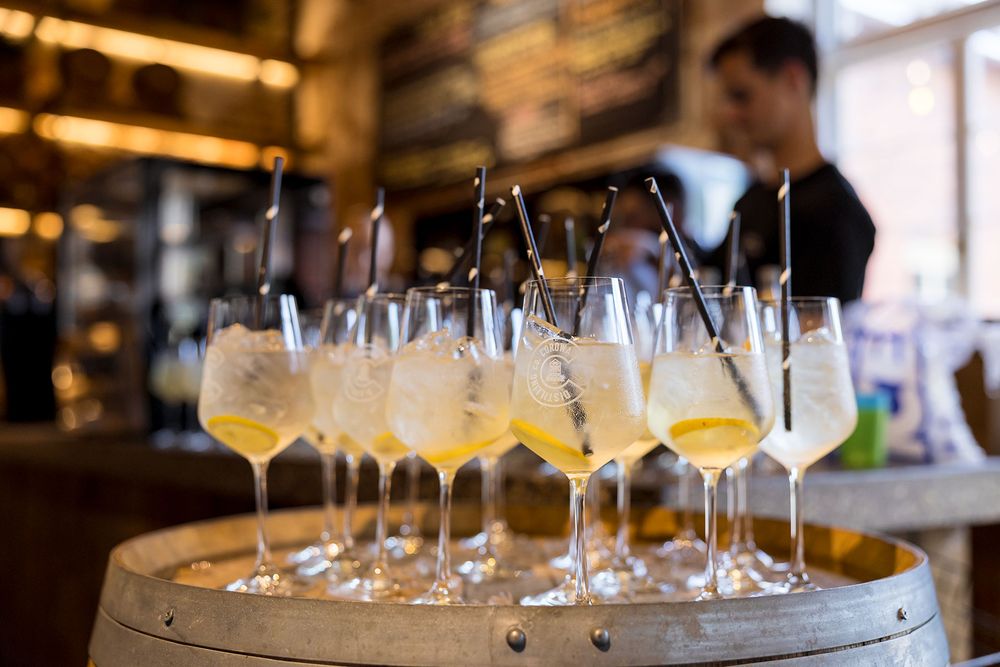 Several glasses of chilled sparkling cocktails with long stirrers sit on a wooden barrel while a bartender stands in the background.