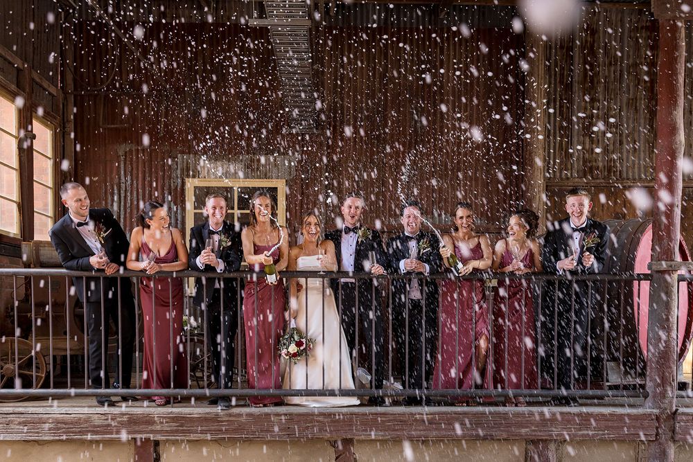 Wedding party in burgundy dresses and dark suits poses on a rustic wooden balcony around the bride spraying champagne.