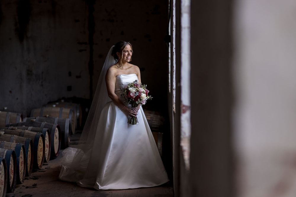 Bride in a strapless white wedding gown and veil holds a bouquet while gazing out a window beside rustic wine barrels at the Corowa Dist.