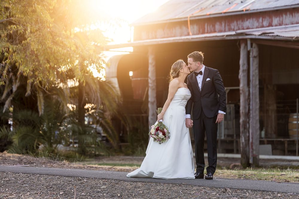 Bride in a white gown holding a bouquet shares a romantic moment with a groom in a black suit outside a rustic barn at golden sunset.