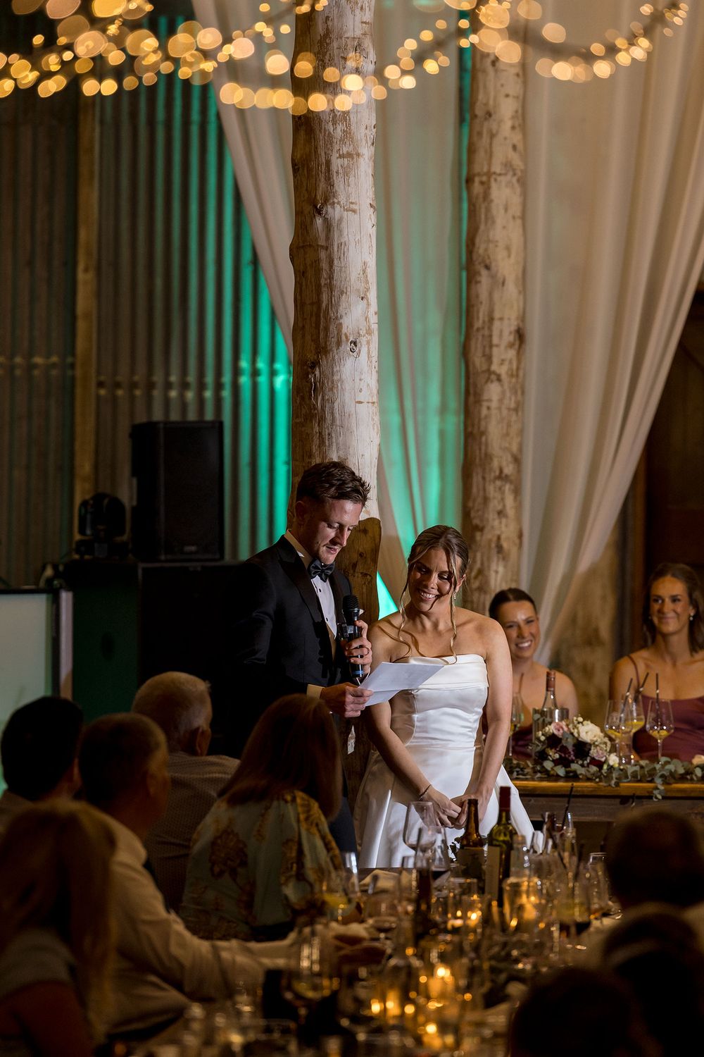 Groom in a tuxedo reads a speech beside his smiling bride in a white gown at a candlelit wedding reception with guests looking on.